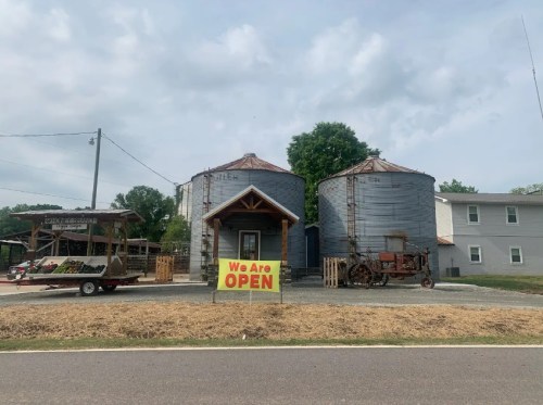 Two large silos with a sign reading "We Are OPEN" in front, alongside a small building and a vintage tractor.