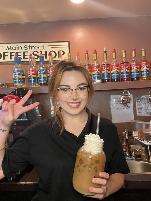 A barista smiles and holds a large iced coffee topped with whipped cream, with colorful syrup bottles in the background.