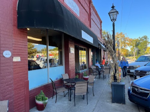 A red brick building with a black awning, outdoor seating, and a lamppost, set in a small-town street.