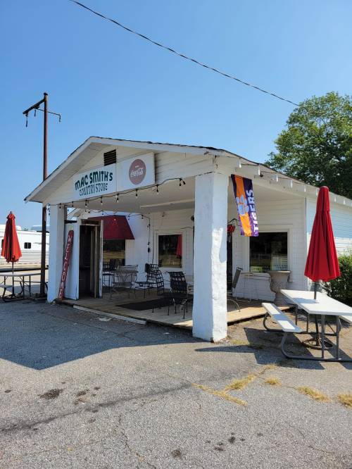 A small, white building with a sign reading "Mac Smith's Country Store," featuring outdoor seating and red umbrellas.