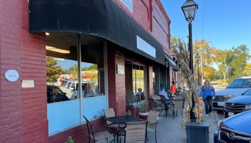 A red brick building with a black awning, outdoor seating, and people walking by on a sunny day.