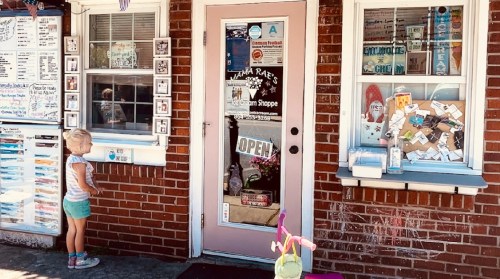 A young child stands by the entrance of a quaint shop with a pink door, surrounded by colorful signs and decorations.
