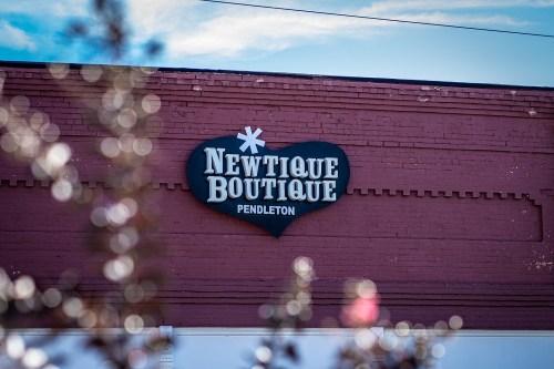 Sign for Newtique Boutique in Pendleton, displayed on a red brick wall with a blue sky in the background.