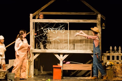 A stage scene with actors performing in a barn setting, featuring a sign that reads "Some Pig" and a character in overalls.