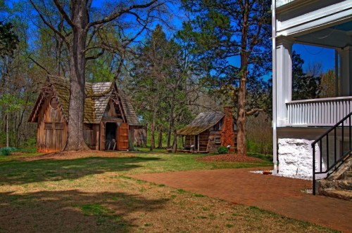 A scenic view of a grassy area featuring two rustic buildings and a large tree under a clear blue sky.