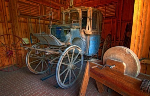 An old wooden carriage inside a rustic barn, surrounded by tools and wooden wheels.