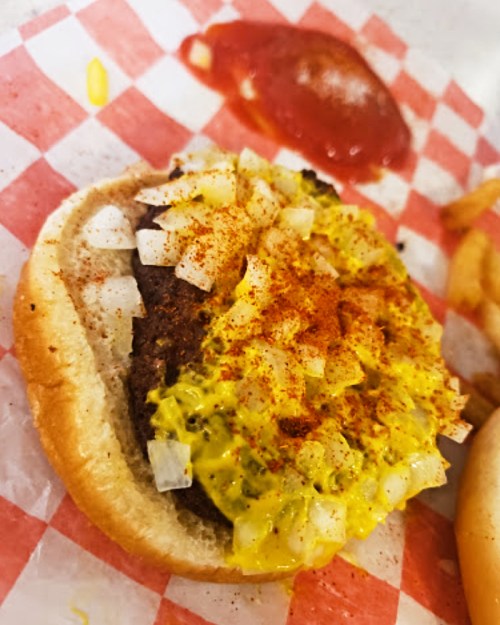Close-up of a hamburger with mustard, diced onions, and seasoning on a checkered paper, with a side of ketchup.