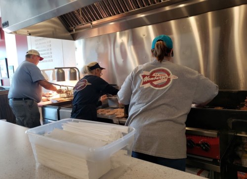 Three cooks prepare food in a busy kitchen, with a container of utensils in the foreground.