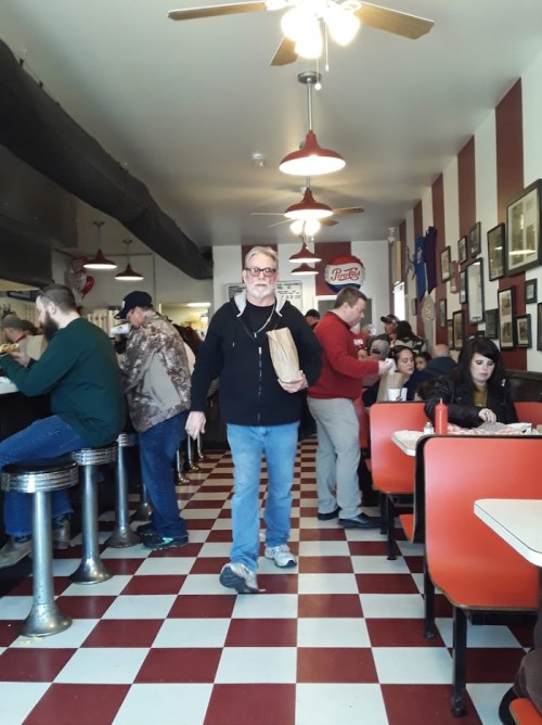 A busy diner with red and white checkered floors, featuring patrons at the counter and tables, and a man walking with a bag.