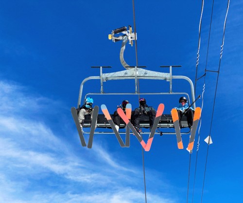 Four skiers on a ski lift against a clear blue sky, with colorful skis hanging below.