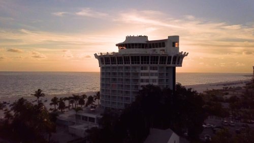 A modern beachfront hotel at sunset, with palm trees in the foreground and the ocean in the background.