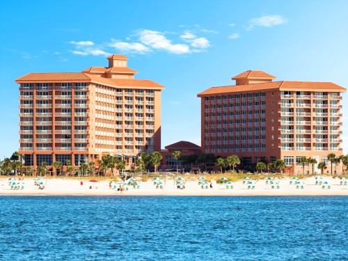 Two large beachfront hotels with orange roofs, surrounded by palm trees and beach chairs, under a clear blue sky.