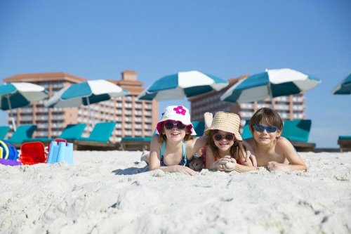 Three children in sunglasses and hats lie on the beach, smiling with umbrellas and lounge chairs in the background.