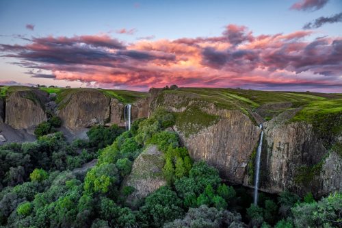 A scenic view of waterfalls cascading down rocky cliffs, surrounded by lush greenery under a colorful sunset sky.