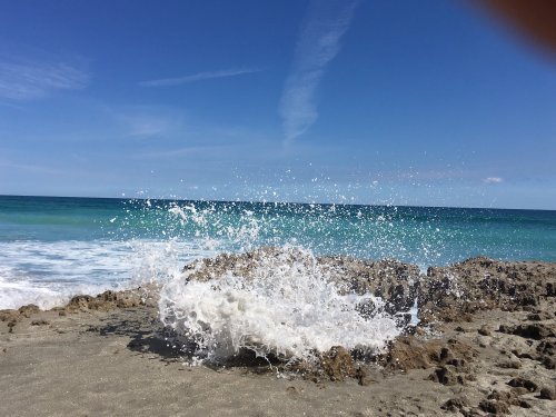 Waves crashing against rocky shore under a clear blue sky with scattered clouds.