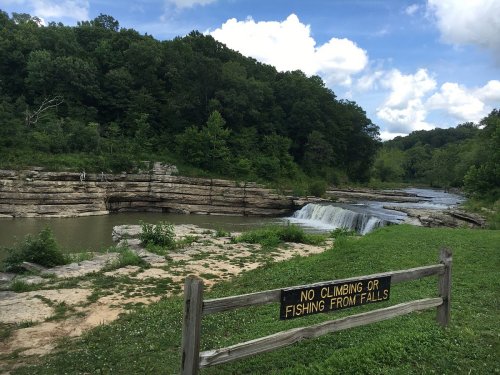 A serene landscape featuring a waterfall and a river, with a sign warning against climbing or fishing from the falls.
