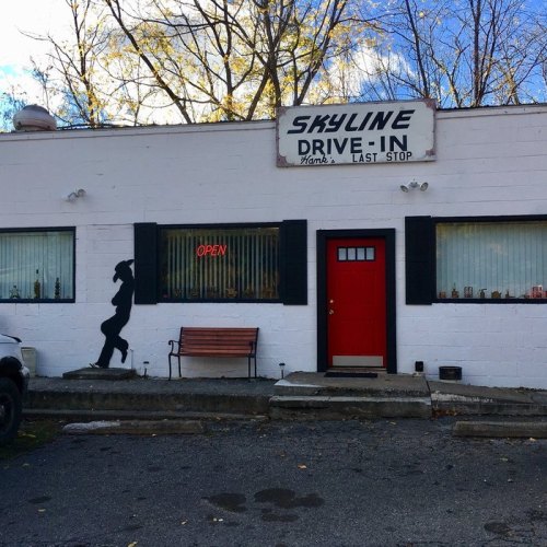 A retro drive-in diner with a red door, open sign, and a silhouette of a person on the wall. Trees in the background.
