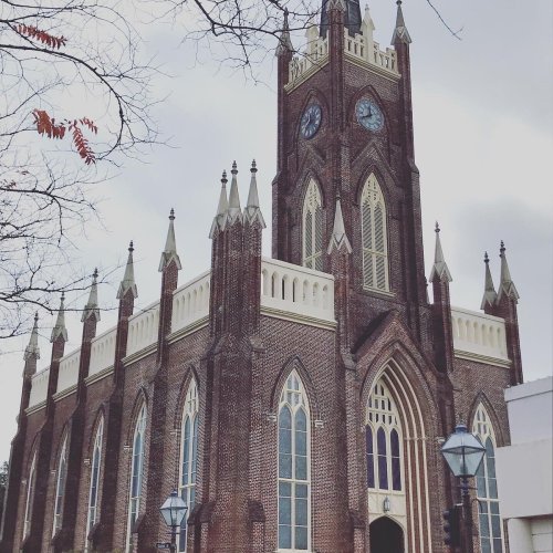 A tall, brick church with a clock tower and pointed spires, surrounded by bare trees and a cloudy sky.