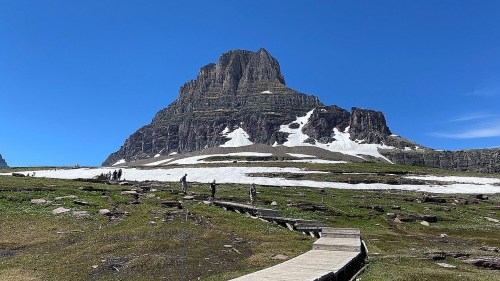 A scenic mountain landscape with a prominent peak, snow patches, and a wooden path leading through green terrain.
