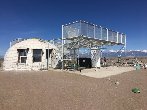 A modern dome structure with a metal frame and a rooftop deck, set against a clear blue sky and distant mountains.