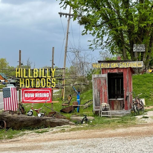 A rustic gas station sign with "Hillbilly Hotdogs" and "Now Hiring," surrounded by vintage items and greenery.