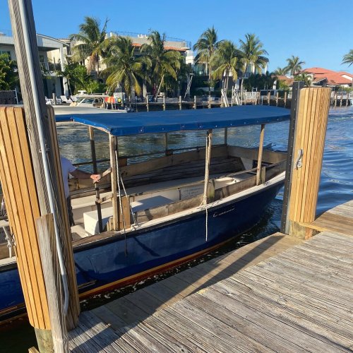 A blue boat docked at a wooden pier with palm trees and waterfront homes in the background under a clear blue sky.