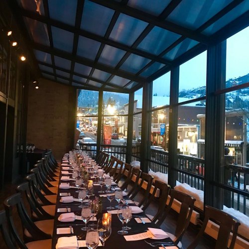 A long dining table set for a meal, with a view of a snowy street and mountains through large glass windows.