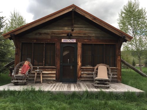 A rustic wooden cabin with a porch, featuring rocking chairs and surrounded by lush green grass and trees.