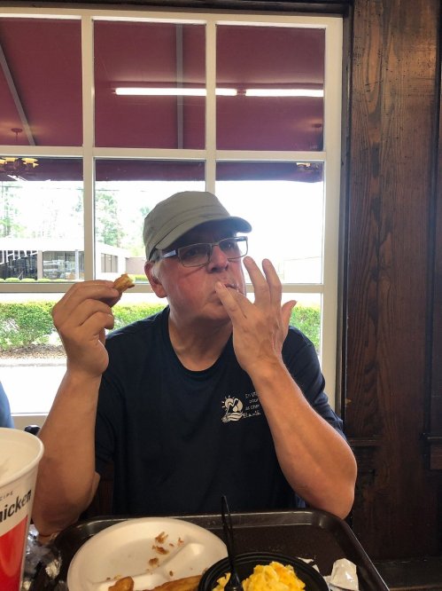 A man in a cap enjoys a meal, licking his fingers while seated at a table in a casual dining setting.