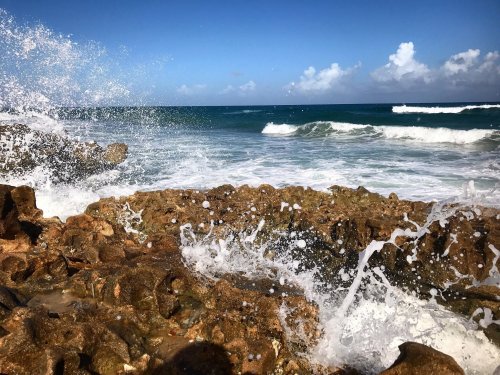 Waves crashing against rocky shore under a clear blue sky with scattered clouds.