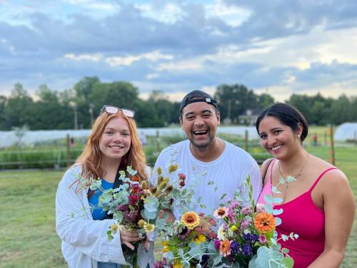 Three smiling individuals hold colorful flower bouquets in a lush outdoor setting with a cloudy sky.