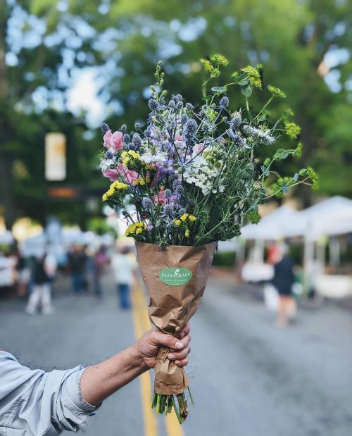 A hand holds a bouquet of colorful wildflowers wrapped in brown paper, with a market scene in the background.