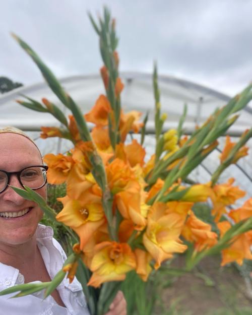 A person with glasses smiles while holding a large bouquet of orange and yellow gladiolus flowers in a greenhouse.