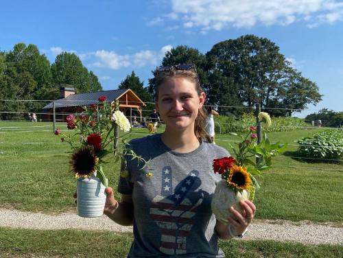 A woman holds two flower arrangements outdoors, smiling in front of a green field and a wooden structure.
