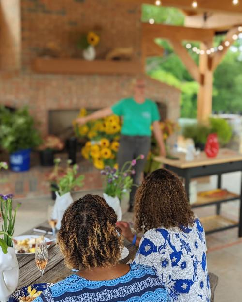 Two women with curly hair sit at a table, enjoying a meal outdoors, while a speaker presents in the background.