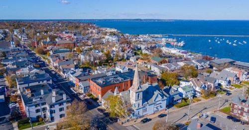 Aerial view of downtown Plymouth, MA, showing historic architecture, a church spire, and boats in the harbor.