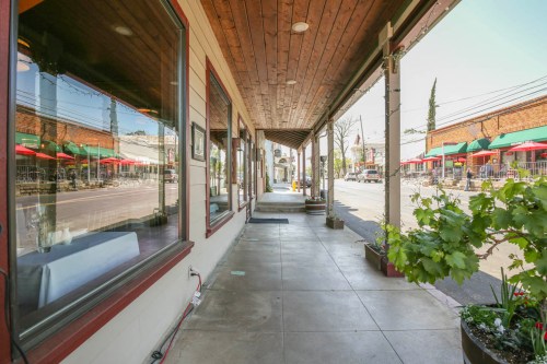 A sidewalk view of a restaurant with outdoor seating, featuring red umbrellas and a tree-lined street in the background.