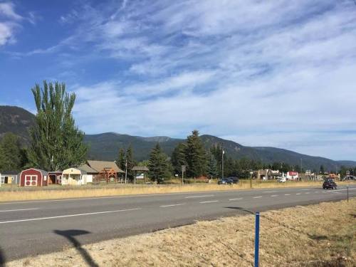 A rural road lined with houses and trees, with mountains in the background under a partly cloudy sky.