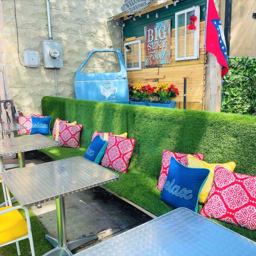 Colorful outdoor seating area with green turf, patterned cushions, and a food stand in the background.