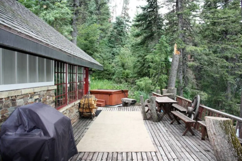 A wooden deck surrounded by trees, featuring a hot tub, grill, and rustic furniture.