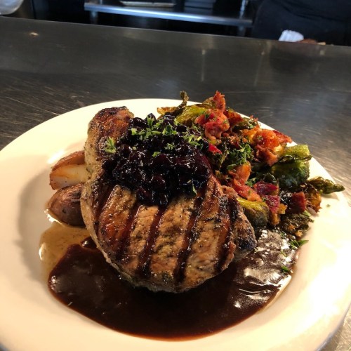 Grilled pork chop with a berry sauce, served with roasted potatoes and sautéed greens on a white plate.