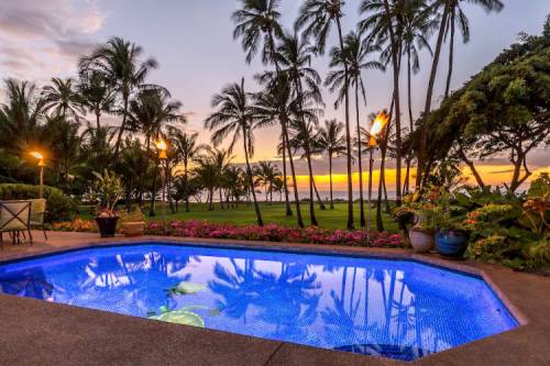 A serene pool surrounded by tropical plants and palm trees, with a sunset over the ocean in the background.