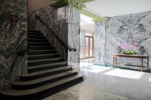 A marble-finished interior with a staircase, large windows, and a table featuring potted orchids.