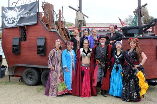 A group of performers in colorful costumes poses in front of a pirate-themed ship at an outdoor event.