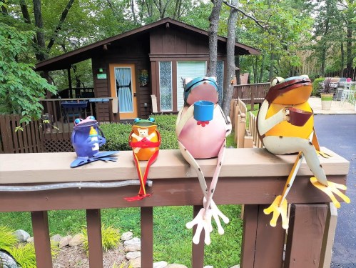 Four colorful frog sculptures sit on a wooden railing, with a cabin and trees in the background.