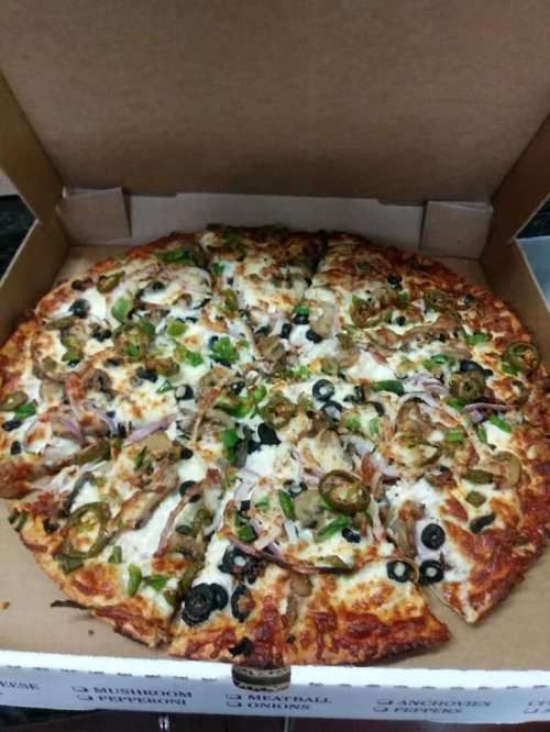 A pizza in a box, topped with cheese, olives, green peppers, onions, and jalapeños, sliced into eight pieces.