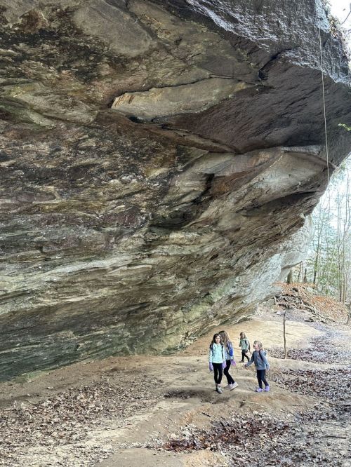 Three people walking under a large rock overhang in a wooded area, with fallen leaves on the ground.