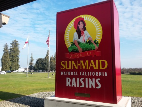 Sign for Sun-Maid Natural California Raisins, featuring a woman with grapes, with flags and trees in the background.
