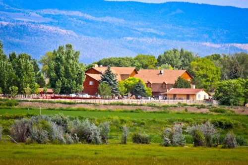 A scenic view of a ranch house surrounded by trees and mountains, with green fields in the foreground.