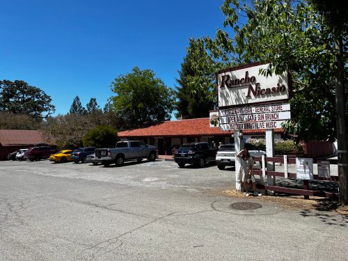 A sunny view of Rancho Nicasio, featuring a restaurant and parking lot with cars, surrounded by trees.